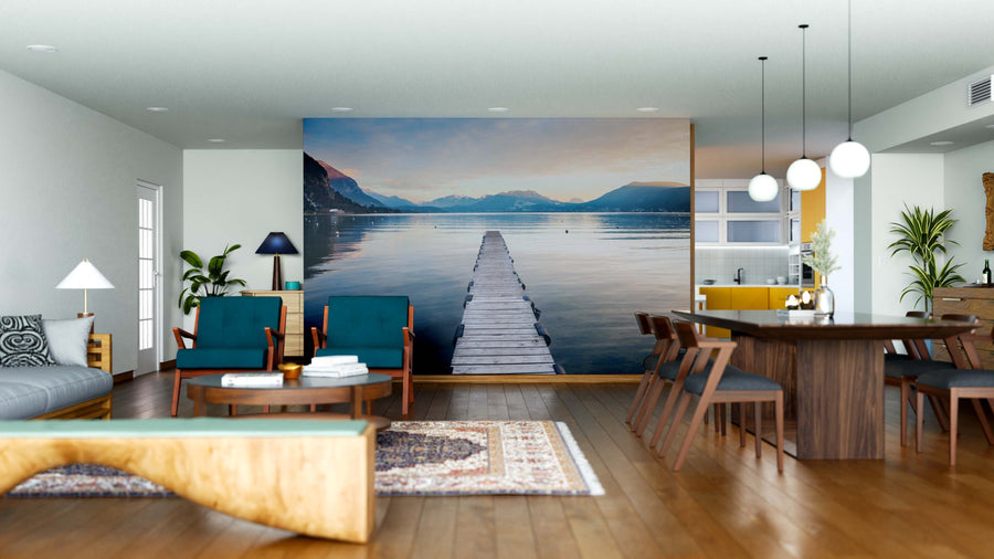 Modern open-plan living and dining room with wood floors, teal and wood furniture, rugs, pendant lights, and plants. The Path to Serenity Wall Mural features a dock on a calm lake at sunset with distant mountains, creating an inviting space.