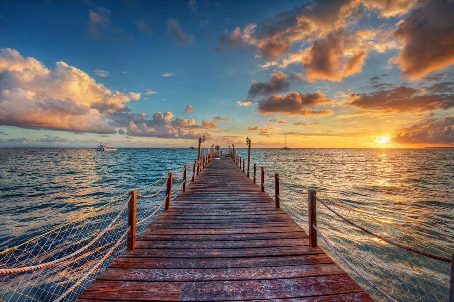 A modern open-plan living and dining room has wooden furniture, blue chairs, pendant lights, and a large Peaceful Ocean View Wall Mural showing a wooden pier stretching into a serene sea at sunset with dramatic clouds and golden reflections.