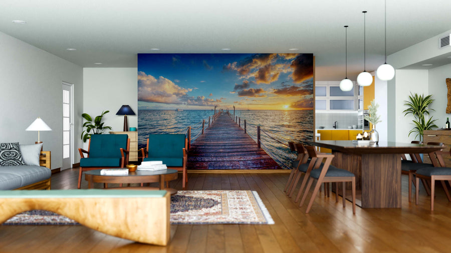 A modern open-plan living and dining room has wooden furniture, blue chairs, pendant lights, and a large Peaceful Ocean View Wall Mural showing a wooden pier stretching into a serene sea at sunset with dramatic clouds and golden reflections.