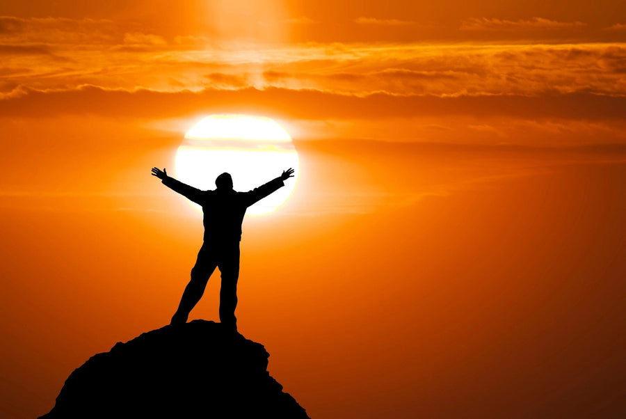 A modern living-dining room with wooden floors, dark blue chairs, and a large Peak of Mountain Wall Mural featuring a person standing triumphantly on a mountain at sunset with orange clouds and a glowing sun.