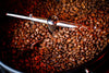 Close-up of roasted coffee beans swirling in a metal roasting machine, captured in warm, rich tones—Perfect Roast Wall Mural brings café vibes to your space as a custom printed or peel and stick mural.