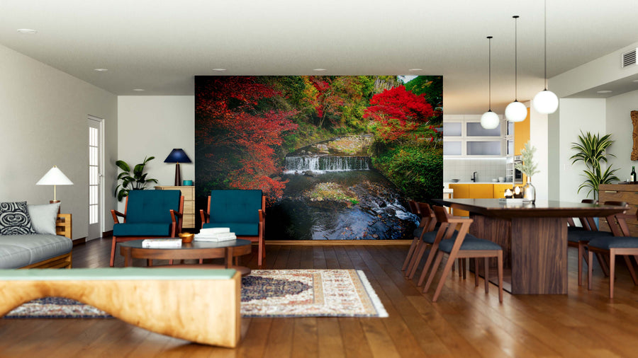 A modern open-plan living and dining area with wood flooring, teal chairs, a wooden table, and a kitchen in the background. The focal wall features the Picturesque Autumn Scene Wall Mural showcasing a forest waterfall.