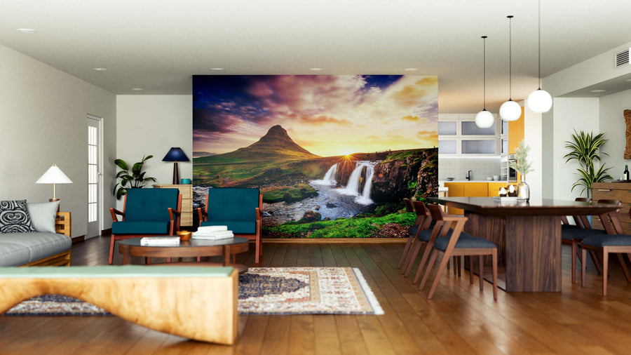 A modern open-plan living and dining area with wooden floors, blue and wood furniture, pendant lights, and a striking Picturesque Sunset Wall Mural depicting mountains, waterfalls, and a vibrant sky. The space is bright with natural and artificial light.