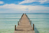 The Pier 8 Wall Mural depicts a wooden pier stretching into a serene, turquoise sea under a blue sky with scattered clouds, transitioning to deep blue farther out for a tranquil, summery feel.