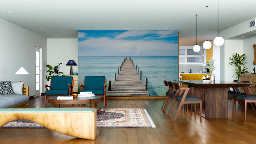 A modern open-concept living and dining area features wood furniture, blue chairs, pendant lights, and the Pier Escape Wall Mural—a custom peel-and-stick mural of a wooden pier over calm sea—adding depth and tranquility to the bright space.