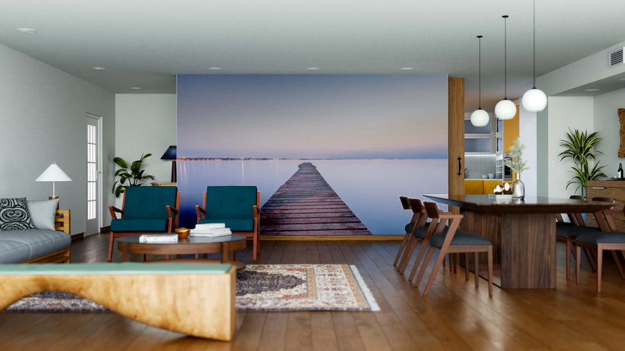 A modern living-dining area with wood floors, blue chairs, and a rug features the Garda Lake Wall Mural—a peel and stick design of a dock reaching into calm water under a pastel sky—creating a serene focal point in the open space.