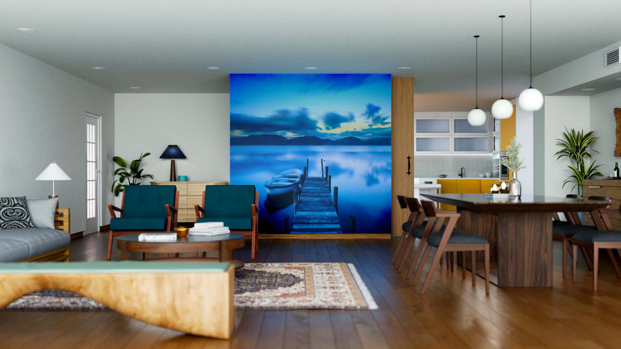 A modern open-concept living and dining room showcases mid-century furniture, wooden floors, and the Jetty and Boat Wall Mural, depicting a tranquil dock at sunset. Cozy rugs, lush plants, and pendant lights complete the inviting atmosphere.