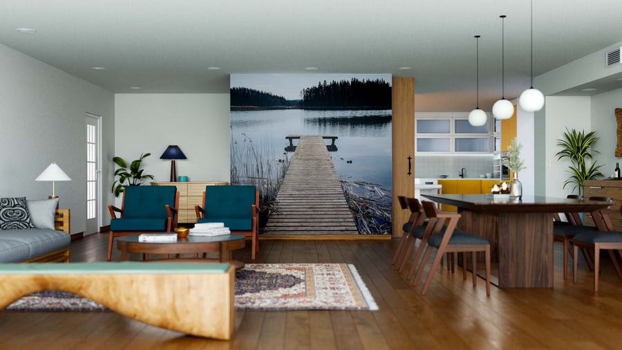 A modern open-plan living and dining room with wooden floors, teal chairs, a patterned rug, and a large Lakeside Serenity Wall Mural of a dock leading into a tranquil lake. The dining area includes wooden chairs and pendant lights over the table.