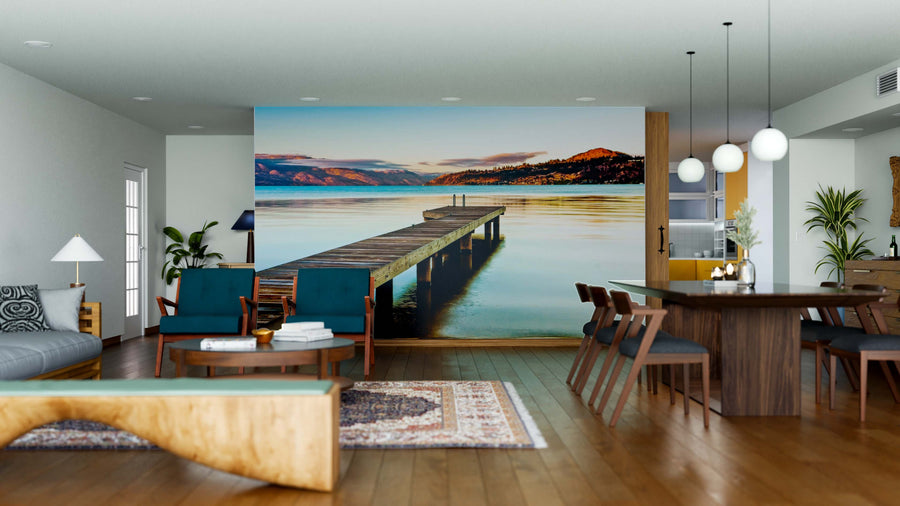 A modern living-dining space with wooden floors showcases the Pier and Beautiful Lake Wall Mural, featuring a dock at sunset. Contemporary furniture, a rug, plants, and pendant lights add a cozy, stylish touch.