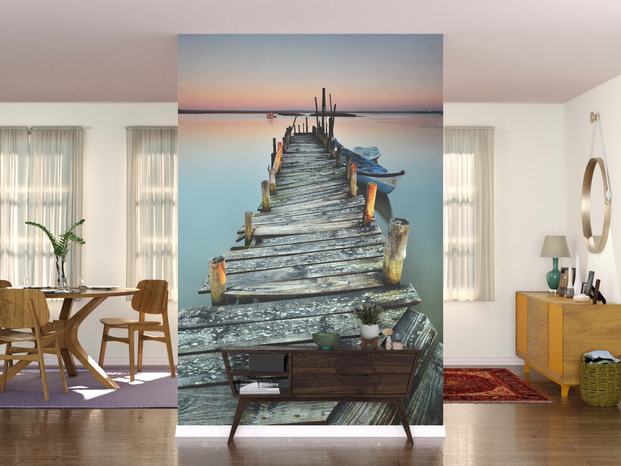 A modern living space highlights the Rustic Dock Wall Mural, showing a wooden pier at sunset with a boat. Wooden furniture, greenery, and a round mirror enhance the cozy vibe and complement the mural’s tranquil setting.