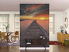 A modern living room showcases the Serenity at Dusk Wall Mural, featuring a wooden pier at sunset. Light walls, wooden furniture, a round mirror, plants, and sheer curtains complete the warm and inviting space.