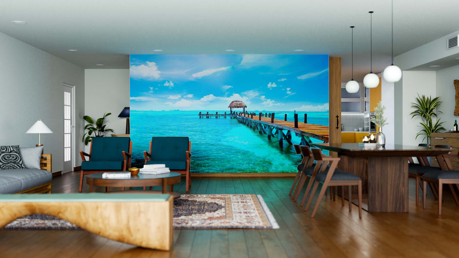 Modern living and dining room with wooden furniture and blue chairs. The Tidebound Tranquility Wall Mural displays a turquoise ocean with a long pier, adding scenic depth, while plants and rugs warm the spacious, well-lit interior.