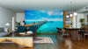 Modern living and dining area with wood floors and mid-century furniture; the Tropical Horizon Wall Mural features a vivid tropical pier extending into turquoise water under a sunny sky, creating an ocean view illusion indoors.