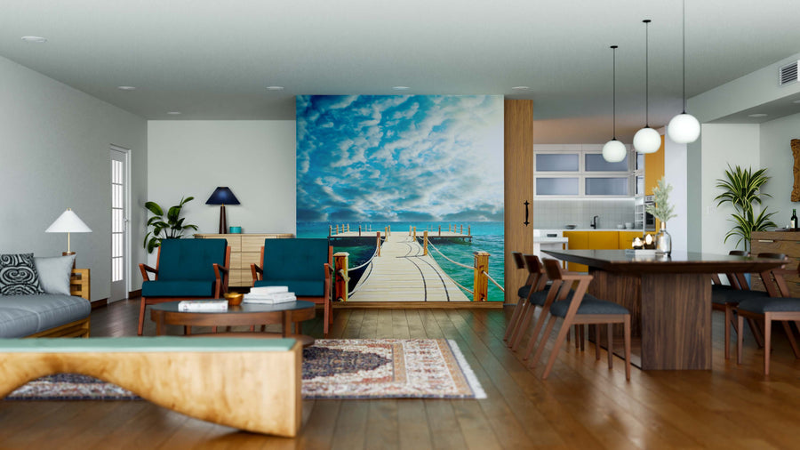 A modern open-plan living and dining room with wooden floors, teal and wood furniture, and a kitchen features the Tropical Wooden Pier Wall Mural—a vibrant peel-and-stick mural showing a pier leading to the ocean under a blue sky.