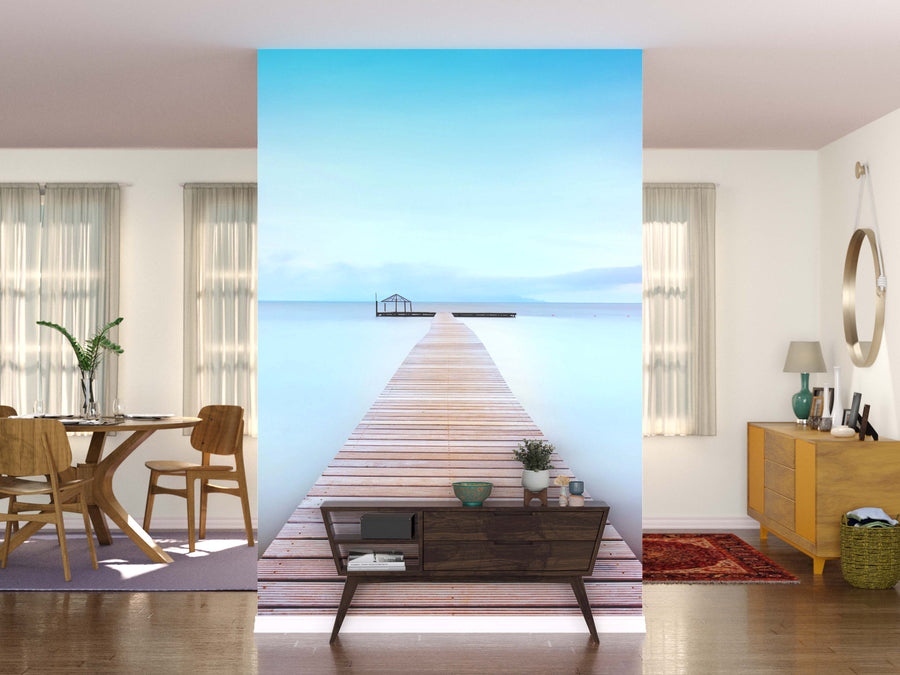 A modern living space features the Tuscan Coast Wall Mural divider showing a wooden pier over calm blue water. One side holds a dining table and chairs; the other, a dresser, mirror, and lamp. Large windows with light curtains brighten the room.