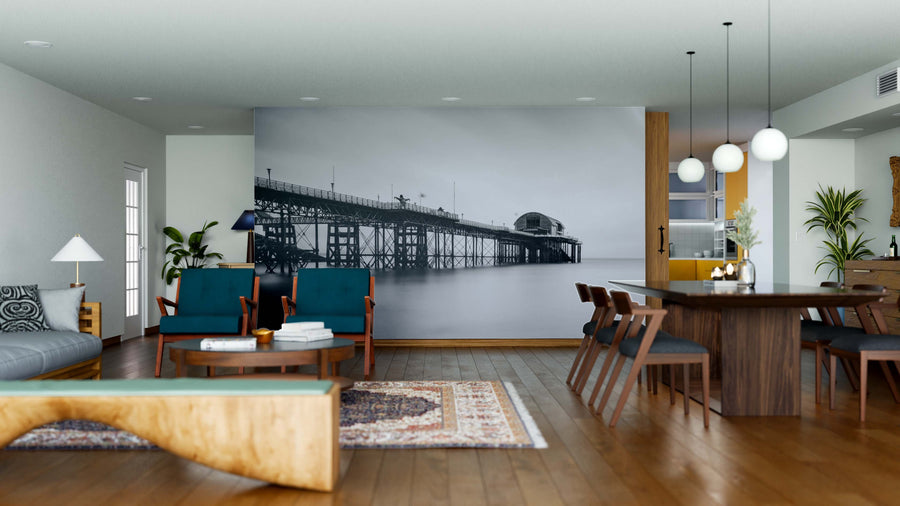 Modern living and dining area with wooden floors, mid-century furniture, teal chairs, round tables, hanging lights, and a Victorian Pier Wall Mural featuring a pier over water. Antique and contemporary decor add to the inviting atmosphere.
