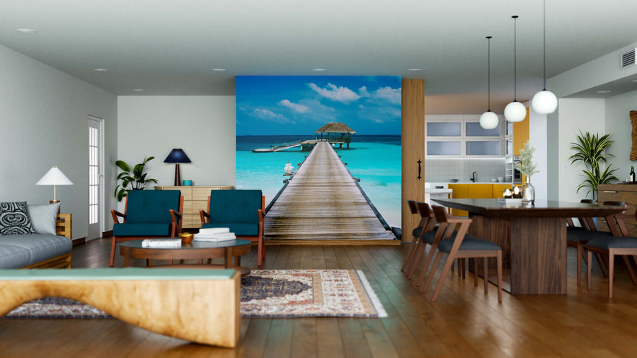 A modern, open-plan living and dining room with wood floors, teal chairs, and a rug features the Water Bungalow Wall Mural—a peel-and-stick mural of a pier over turquoise water for a beach escape feel. The kitchen is visible on the right.