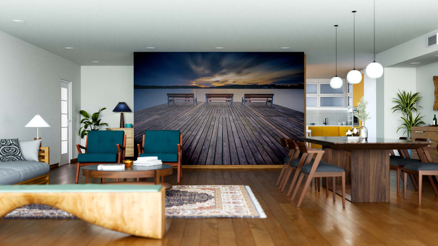 A modern living and dining room features wood furniture, teal chairs, a rug, and the Pier and Benches Wall Mural showing a dock at sunset for an airy feel. Pendant lights hang above the dining table.