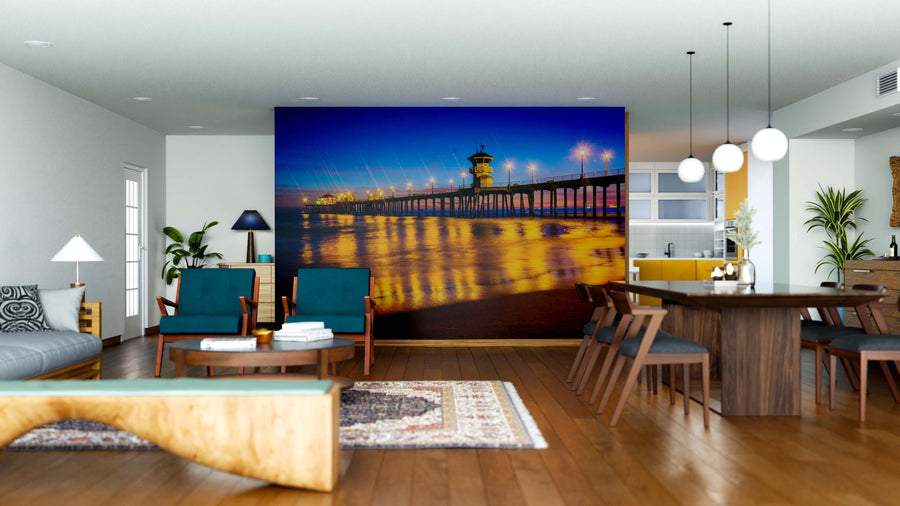 A modern open living and dining space features wood furniture, rugs, pendant lights, and the Pier at Night Wall Mural—a vivid depiction of a brightly lit pier over water—creating a striking focal point in the room.