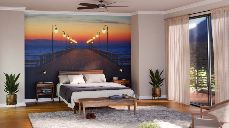 A modern bedroom includes a bed with gray bedding, wooden nightstands, plants, a wooden bench, and a large Pier in Sunrise Wall Mural. Sliding glass doors with sheer curtains open to a balcony with greenery outside.
