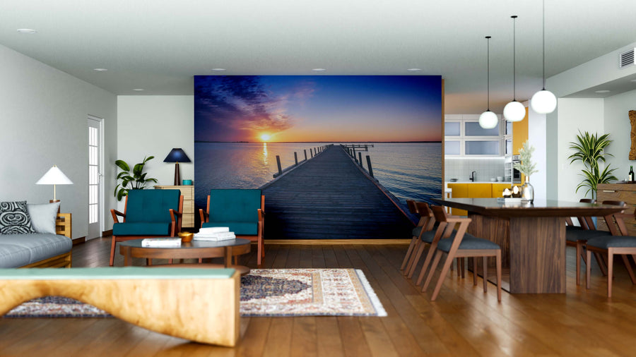 A modern open-plan living and dining room features wood furniture, blue armchairs, rugs, and the Pier to Tranquility Wall Mural—a striking image of a dock at sunset—bringing depth and calm to the contemporary space.