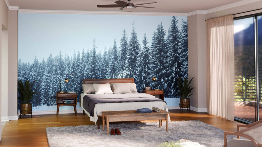 A modern bedroom showcases the Pine Trees Forest Covered with Snow Wall Mural behind a wooden bed, flanked by nightstands and lamps, with a bench at the foot, plants, a soft rug on the wooden floor, and sheer curtains by the glass door.