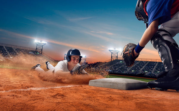 The Players Infield Wall Mural features a baseball player in a white uniform and helmet sliding headfirst toward home plate, kicking up dirt under stadium lights at sunset, captured on a matte canvas textured peel and stick mural.