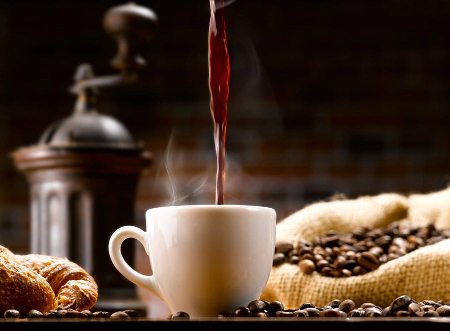 A modern coffee shop with wooden tables, stools, potted plants, and large windows features the Pouring Fresh Coffee Wall Mural on the back wall, depicting a coffee cup being filled with beans and a grinder for a warm, inviting vibe.
