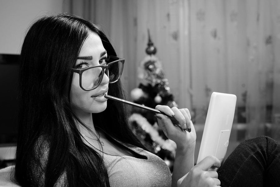 A woman with long dark hair and large glasses is seated indoors, holding a tablet and playfully biting a pen. Behind her, the blurred scene resembles the Pretty Girl in Glasses Wall Mural, with a Christmas tree adding to the ambiance. She wears a fitted top and gazes directly at the camera in this black-and-white image.