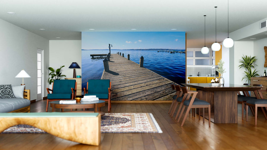 A modern living-dining area with wood floors, blue chairs, and a long dining table is enhanced by the Public Dock at Marine Point Wall Mural, showcasing a realistic dock over calm water beneath a clear sky.