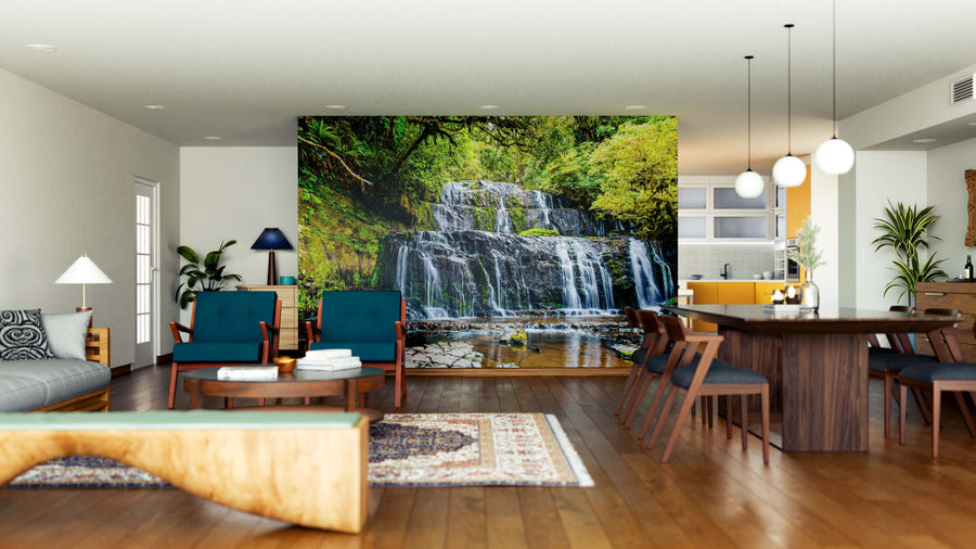 Modern open living-dining area with wooden floors, teal chairs, a rug, and pendant lights. The Purakaunui Falls Wall Mural of a lush waterfall creates a striking focal point. The kitchen in the background features yellow accents.