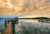 Transform your space with the Quiet Pier Wall Mural, capturing a serene lake scene at sunrise. A wooden pier and covered gazebo, mirrored by soft orange and gold clouds, offer tranquility as tree-lined shores and a lone duck embody peace.