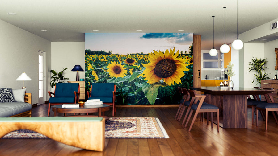 A modern open-plan living and dining room has wooden floors, dark wood furniture, cozy seating, and a Quiet Sunset Blooms Wall Mural with vibrant sunflowers. Pendant lights hang above the dining table, and houseplants provide greenery.
