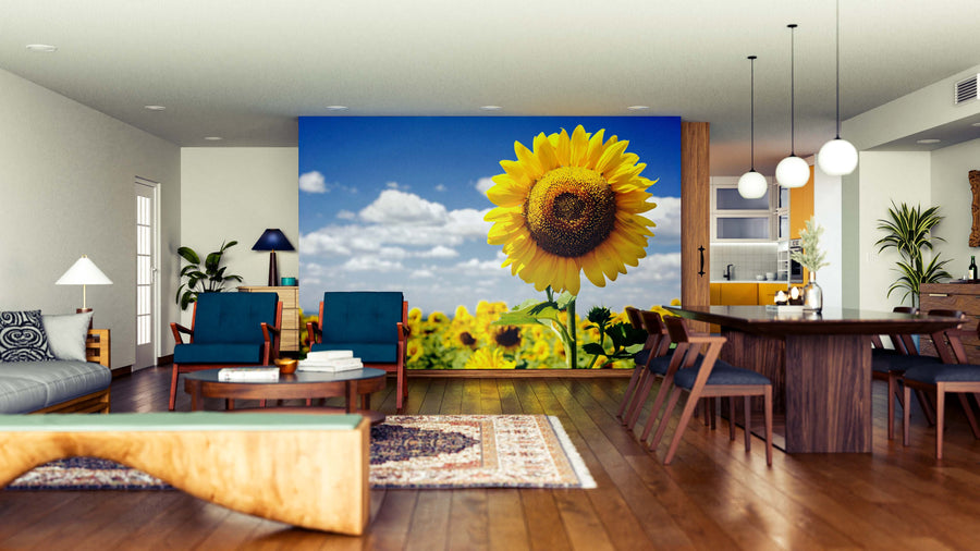 A modern open-plan living and dining room with wooden floors, blue armchairs, and wood furniture is enhanced by the Radiant Summer Flower Wall Mural, bringing a sunflower field under blue sky to the space with pendant lights and patterned rugs.