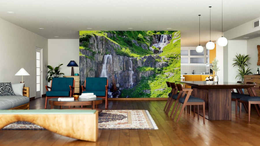 A modern open-plan living and dining room with wooden floors, teal and wood furniture, and pendant lights features the Rapid Waterfalls Wall Mural—a lush, green cliff with waterfalls that serves as a vibrant natural focal point.