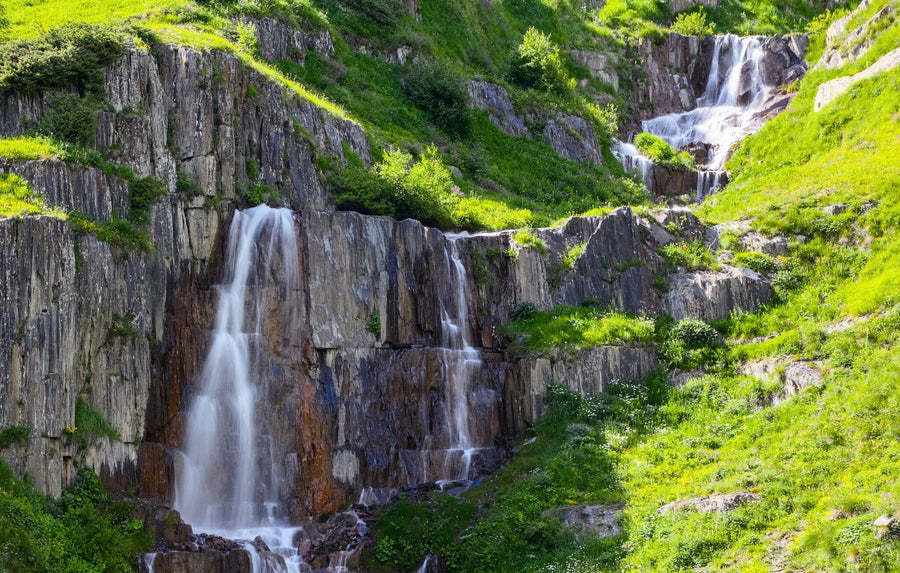 A modern open-plan living and dining room with wooden floors, teal and wood furniture, and pendant lights features the Rapid Waterfalls Wall Mural—a lush, green cliff with waterfalls that serves as a vibrant natural focal point.