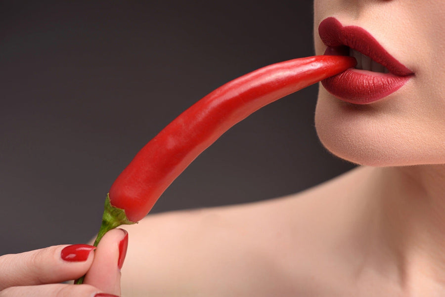 A woman's mouth with bright red lipstick bites a chili pepper, her manicured nails matching the vibrant hues of the Sexy 2 Wall Mural. A dark background contrasts with her light skin, enhancing the striking reds.
