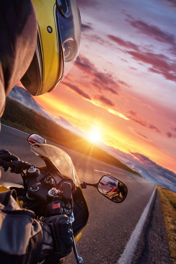 Wearing a yellow helmet, a motorcyclist navigates a winding road at sunset, as the sky resembles the vibrant hues of the Rider at Sunset Wall Mural by  photo. The sun dips low, capturing both motion and scenic beauty.