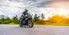 A person rides a black sport motorcycle swiftly along a paved road with blurred greenery, resembling the "Riding into Motion Wall Mural." The rider in black gear and helmet is highlighted by sunlight flares on the right, while clouds fill the sky for added drama.