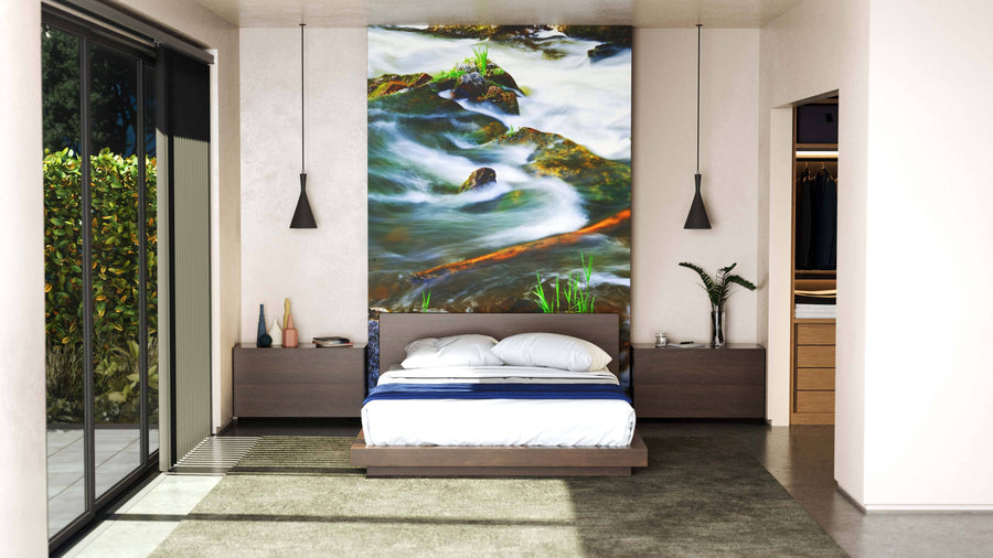 A modern bedroom features a large bed with white bedding and dark wood furniture. Above the bed, the “River Through Woods Wall Mural” adds vibrant color. Pendant lights flank the bed, and sunlight pours in through glass doors on the left.