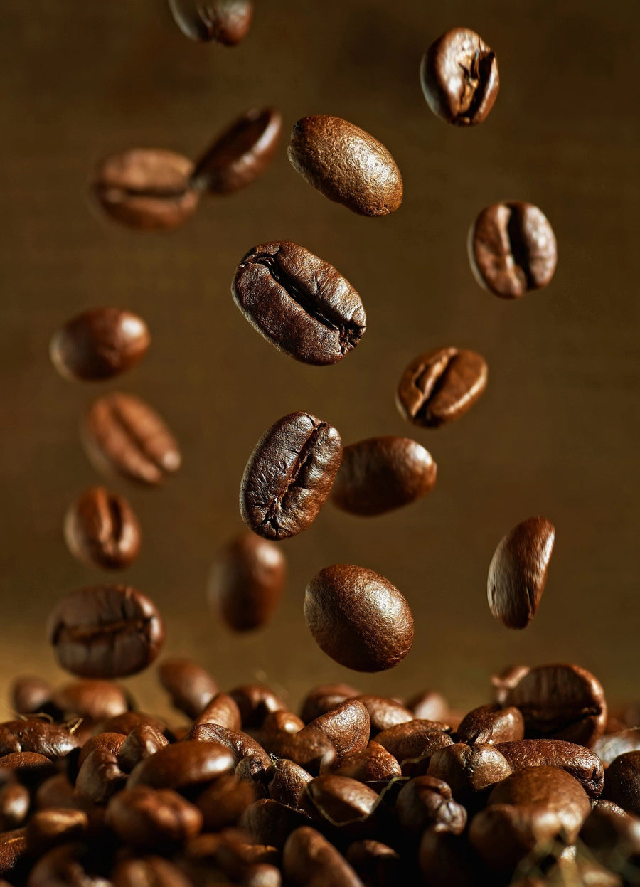 A modern café with wooden tables and stools features the Roasted Coffee Beans Wall Mural, hanging pendant lights, a bulletin board with papers, small potted plants on tables, and large windows that fill the space with natural light.