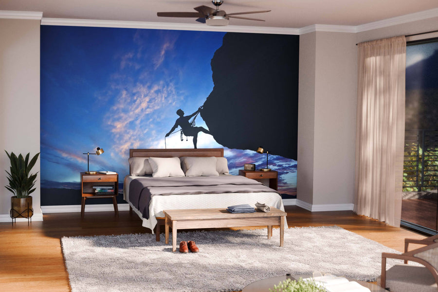 Modern bedroom with a large bed, wooden furniture, and indoor plants. Behind the bed, the Rock Climber Rappelling Wall Mural adds a striking focal point with its silhouette of a climber against a sunset sky.