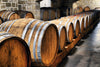 Dimly lit cellar features rows of large wooden barrels with metal bands and polished surfaces, likely for aging wine or spirits, against a stone wall. Custom printed "Wine 17 Wall Mural" by Photo adds depth and charm, while gentle light enhances the rustic atmosphere.