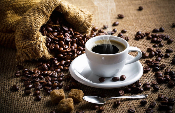A steaming cup of black coffee on a white saucer, with a teaspoon, rests among scattered roasted beans and a burlap sack spilling onto a textured surface. This scene mirrors the earthy tones in our Rustic Roast Wall Mural, with two brown sugar cubes in the foreground.