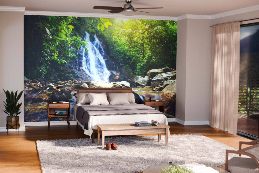 A modern bedroom features the Sai Rung Waterfall Wall Mural behind the bed. Wooden furniture, neutral bedding, a leafy plant, and a beige rug add coziness as sunlight filters through sheer-curtained windows.