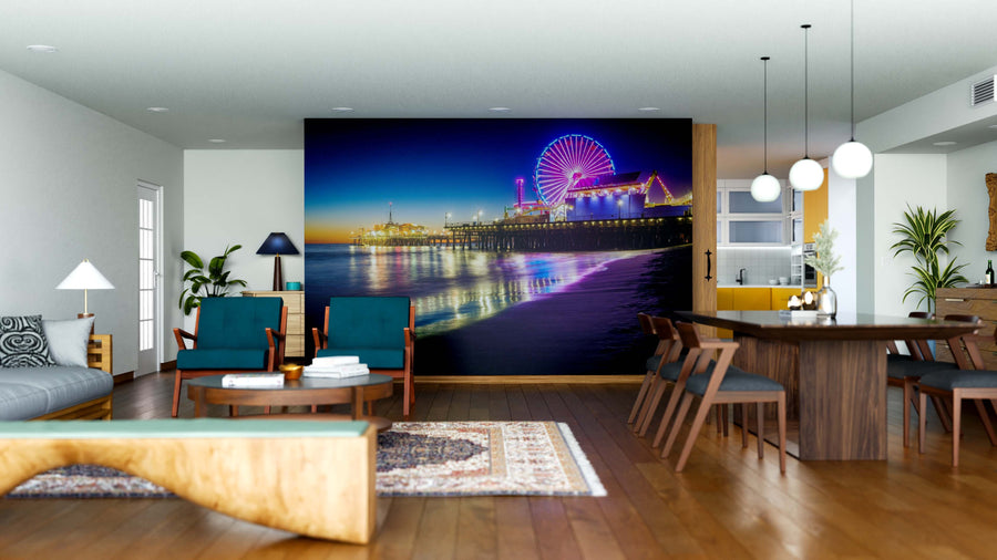 A modern living-dining room features wood furniture and a large Santa Monica Pier at Sunset Wall Mural, with green chairs, a patterned rug, and globe pendant lights.
