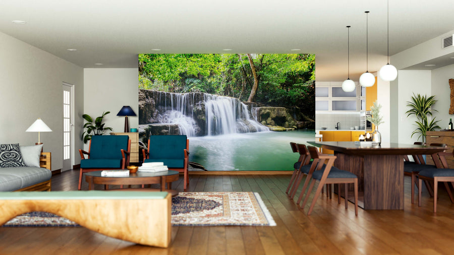 A modern living and dining room with wood floors, teal chairs, a rug, plants, and pendant lights features the Scenic Waterfalls Wall Mural as a calming focal point of lush green forest and waterfall behind the seating area.
