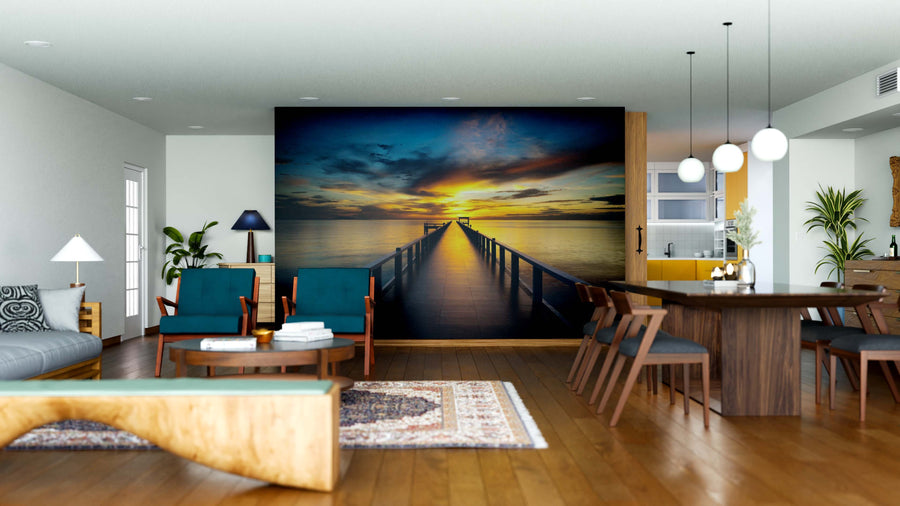 A modern living-dining area with wood furniture and teal chairs features the Sea Bridge Wall Mural, depicting a pier at sunset over water. Soft lighting, plants, a rug, and decor complete the cozy, inviting vibe.