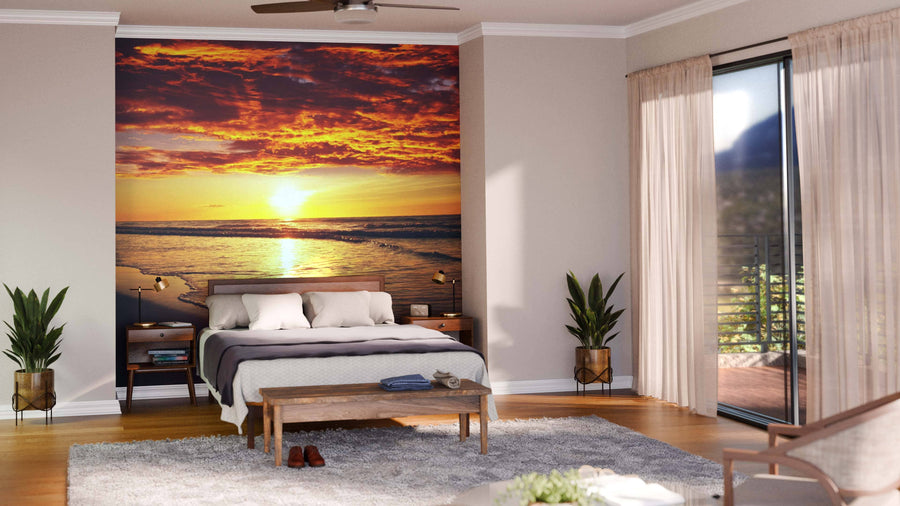 A modern bedroom features the Sea Sunset Wall Mural behind the bed, beige walls, light bedding, two bedside tables, potted plants, a wooden bench, ceiling fan, and sheer curtains over sliding glass doors to a balcony.