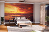 A modern bedroom features a large bed, wooden furniture, and a gray rug. Behind the bed, the Sea Water Wall Mural adds warmth with its orange and yellow ocean sunset design. Sunlight filters in through sheer beige curtains over tall windows.